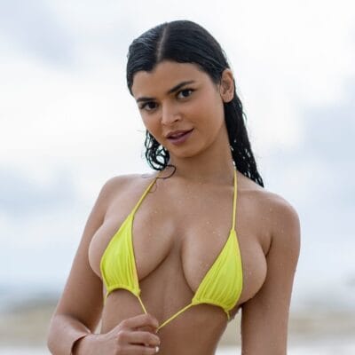 Woman in a neon yellow bikini posing at the beach, facing the camera with a slight smile, wet hair.