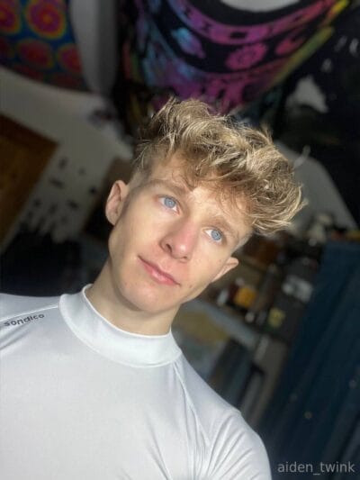 Young man with blond tousled hair and blue eyes wearing a white top, tilting his head in a cluttered room with colorful decor in the background.