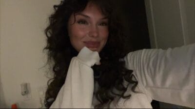 Smiling woman with long dark curly hair wearing a white sweater, posing for a selfie in a dimly lit room.