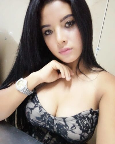 Young woman with long black hair wearing a black lace top and a silver watch, posing with her chin resting on her hand in a neutral setting.] ,