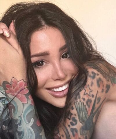 Close-up of a smiling woman with long dark hair and colorful sleeve tattoos on her arm.
