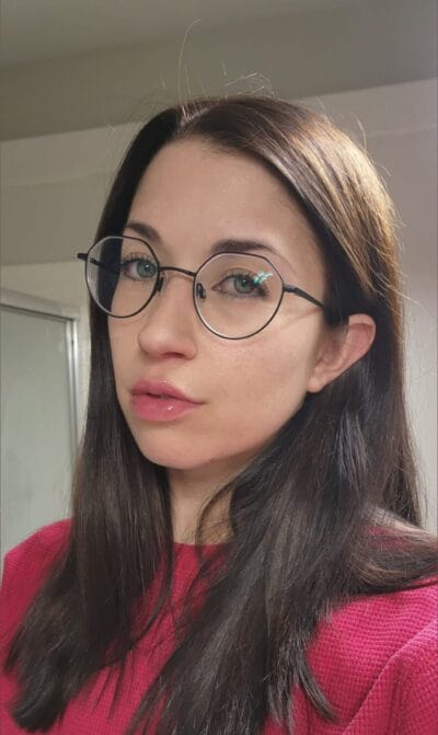 Close-up portrait of a woman with round metal glasses, long dark hair, and a pink knit top, looking at the camera in an indoor setting.