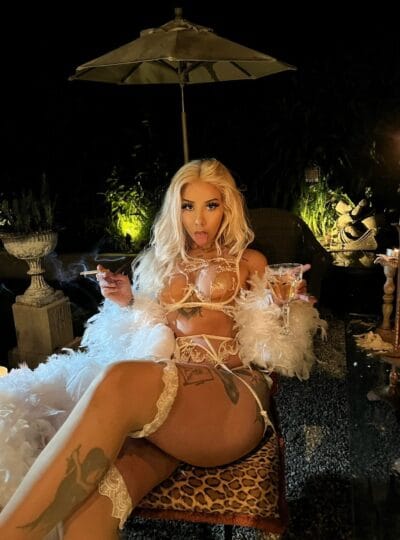 Nighttime outdoor boudoir scene: blonde woman in white lace lingerie and feather boa sits on a leopard-print chair, smoking a cigarette and holding a wine glass under an umbrella. Toward a dimly lit garden backdrop with plants and candles.