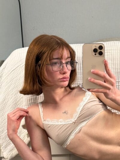 Woman with shoulder-length brown hair and glasses takes a mirror selfie, holding a gold iPhone and wearing a beige lace-trim camisole in a light room center-stage.