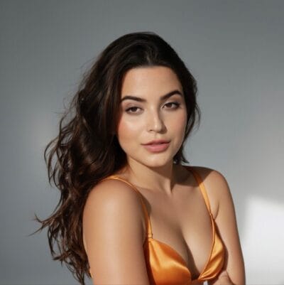 Close-up portrait of a woman with long brown hair wearing an orange satin top, against a gray backdrop.