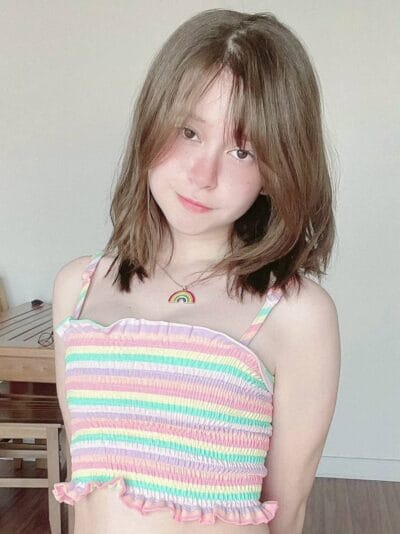 Young woman with shoulder-length brown hair wearing a pastel rainbow-striped crop top and a small rainbow pendant necklace, smiling softly.