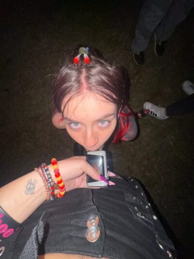 Girl looking up at the camera outdoors at night, wearing colorful bracelets and a beaded hair accessory, holding a small pink phone.