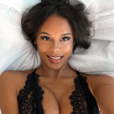 Woman lying on a bed, wearing black lace lingerie, smiling at the camera with makeup and curly hair visible.