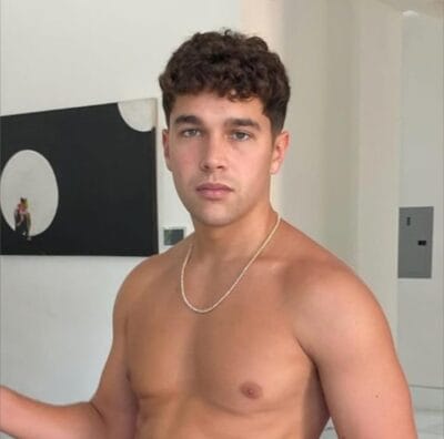 Young man shirtless indoors, wearing a silver chain necklace, facing the camera with neutral expression.