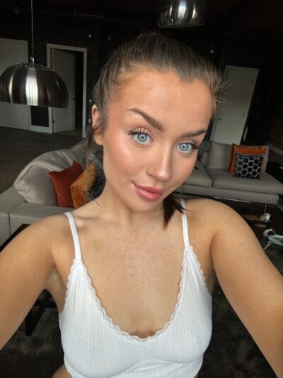 Close-up selfie of a woman with blue eyes and makeup, wearing a white lace top, in a living room with a gray sofa and colorful pillows in the background