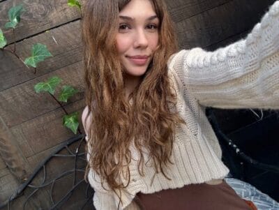 Young woman with wavy brown hair in a cream knit sweater takes a selfie outdoors, standing against a wooden fence with ivy.