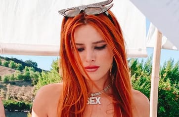 Woman with long red hair wearing sunglasses on her head, looking down outdoors, wearing a necklace that reads 'SEX'.