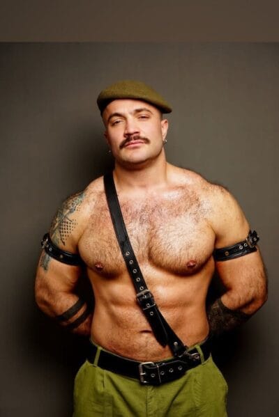 Portrait of a shirtless, tattooed man with a mustache wearing a green beret and black leather harness against a neutral backdrop, facing camera.