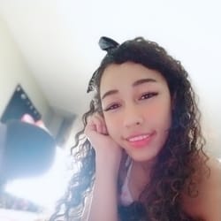 Young woman with curly hair and a black bandana, resting her chin on her hand and smiling at the camera indoors by a bright window.