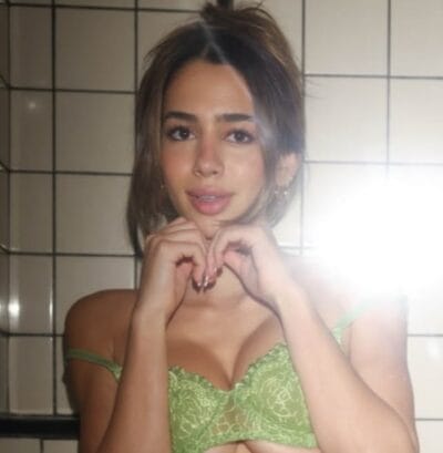 Woman with brown hair wearing a green lace bra, forming a heart shape with her hands in front of a tiled wall.
