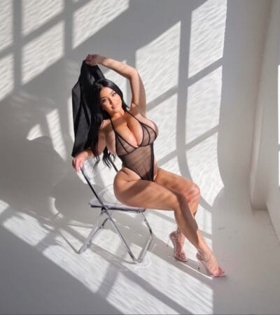 Woman posing in sheer black lingerie on a clear chair, holding a black garment above her head in a sunlit studio.