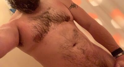 Close-up selfie of a shirtless man showing a hairy chest and arm, with a wristband on the right wrist and a small tattoo on the upper arm.