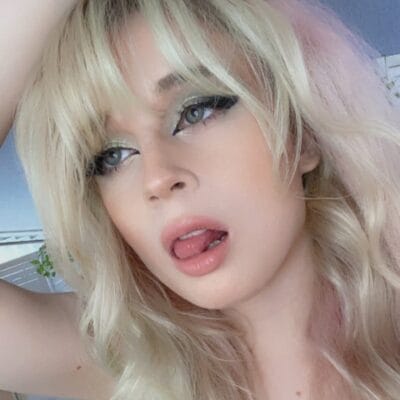 Close-up portrait of a blonde woman making a playful face with lips slightly parted and tongue showing under bold eye makeup.