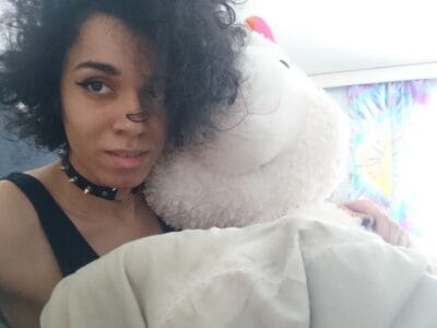 Partial portrait of a person with curly hair, wearing a black sleeveless top and spiked choker, hugging a large white plush toy in a bright room.