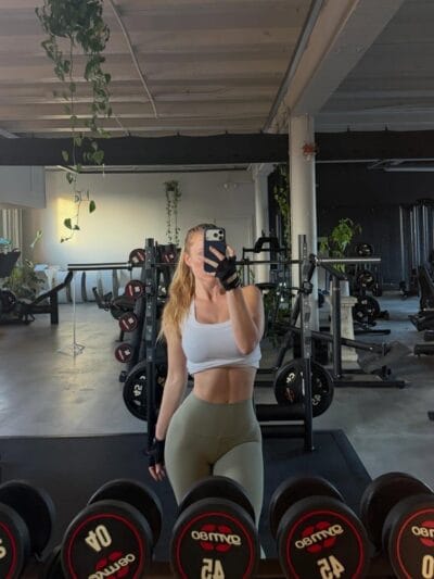 Woman in a white sports bra and olive-green leggings takes a mirror selfie in a gym, wearing fingerless gloves with gym equipment and weights in the background.