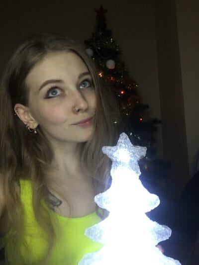 Young woman with blonde hair and a nose ring indoors, beside a glowing ice sculpture with a Christmas tree in the background.