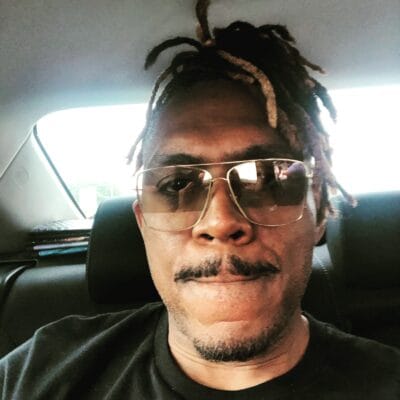 Close-up of a man wearing aviator sunglasses in a car, with dreadlocks visible above his head.
