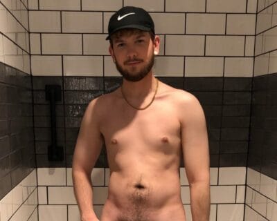 Shirtless man wearing a black Nike cap and gold chain stands in a tiled shower with a handheld showerhead on the wall behind him.