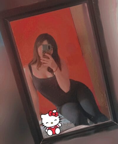 Person taking a mirror selfie in a room with an orange wall, wearing a black tank top and dark leggings; Hello Kitty sticker in the lower corner of the mirror.