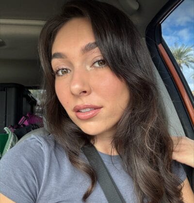 Close-up of a woman with long dark hair wearing a gray shirt and seatbelt in a car, sunny day and palm trees outside the window behind her.