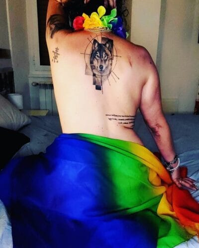 Back view of a person with a wolf tattoo in geometric lines on the upper back, rainbow fabric draped around the hips.