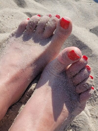 Toes with bright red nail polish partially buried in sandy beach sand.