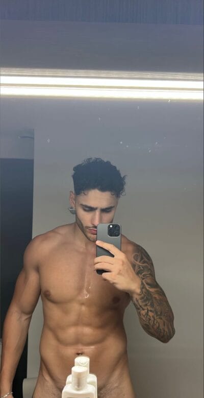 Shirtless man with a tattooed arm taking a mirror selfie in a bathroom, holding a smartphone over his chest, bottles on the sink in front of him.