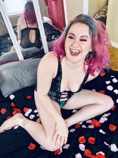 Person with pink and blue hair laughing on a bed in black lingerie, rose petals scattered on a dark sheet; reflection shows back view in a mirror.