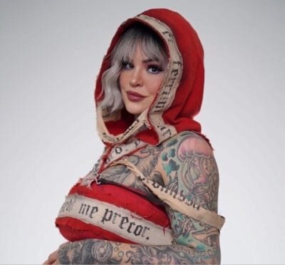 Person with silver hair wearing a red hooded scarf and visible arm tattoos, posing against a light background.