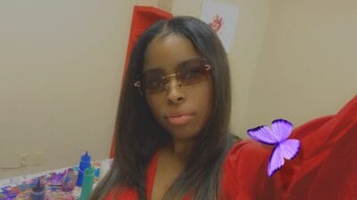 Person in a red outfit wearing tinted sunglasses posing for a selfie in a craft room with paint supplies on the table behind them.