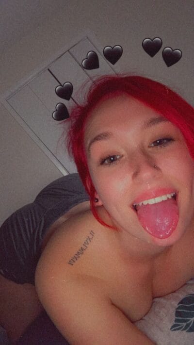 Selfie of a person with bright red hair sticking out their tongue, lying on a bed with black heart stickers around them.