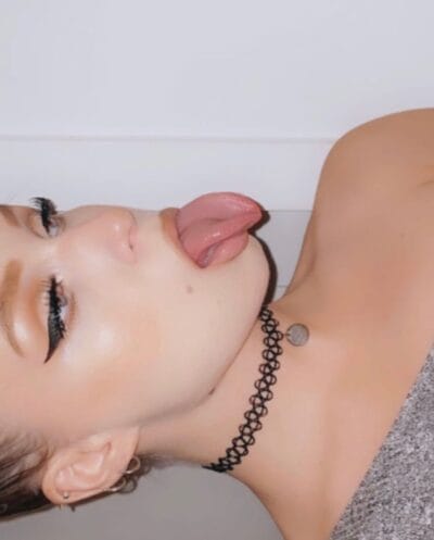 Close-up of a person lying on their side with tongue out, wearing a black spiral choker and a round pendant necklace.