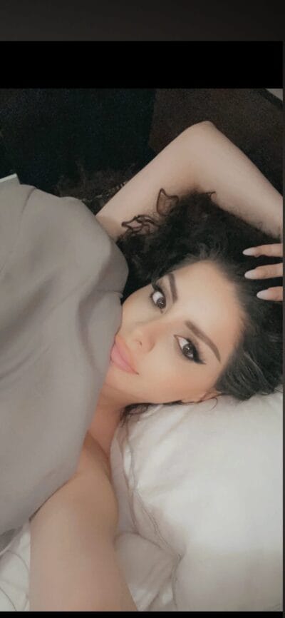 Woman lying on a pillow with a gray blanket, head turned toward the camera, hand resting in her hair, makeup accenting eyes and lips ( tattoo on inner arm visible ).