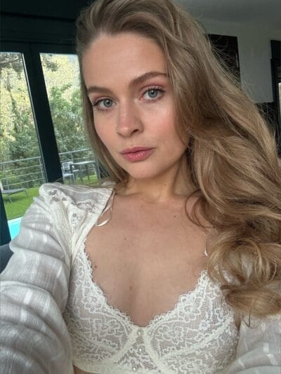 Close-up selfie of a young woman with blue eyes and long wavy blonde hair wearing a white lace top.