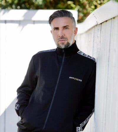 Man leaning against a white wooden fence, wearing a black zip-up jacket with a BRAZZERS logo on the chest.
