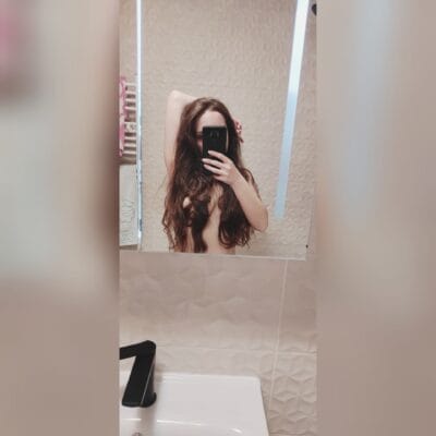 Person with long brown hair taking a mirror selfie in a bathroom, phone obscuring their face.