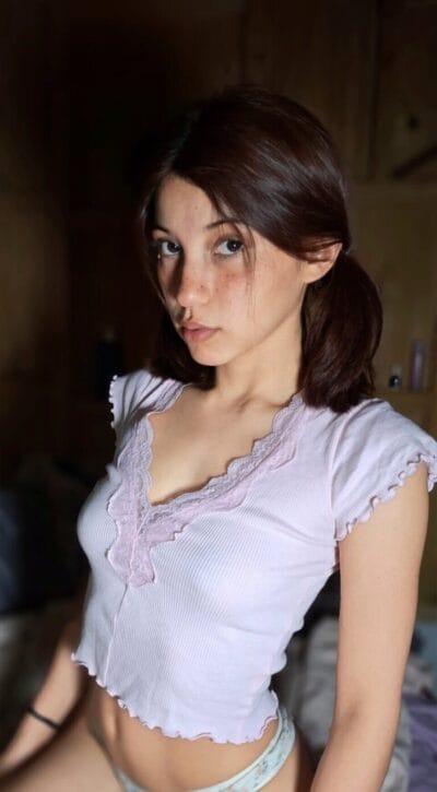 Young woman with dark hair in a light purple lace-trim top, looking at the camera in soft indoor lighting.