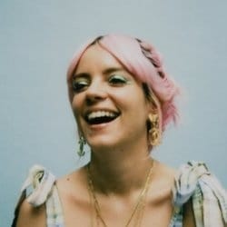 Woman with pink hair laughing, wearing a light blue top with tied shoulder bows and gold jewelry against a pale blue background.