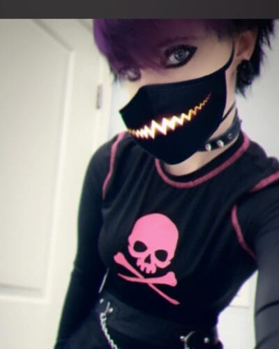 Person wearing a black mask with a yellow zigzag pattern, purple hair, a pink skull shirt, and a black choker on a selfie. (informative fashion photo)