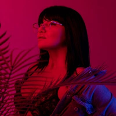 Close-up profile of a person with dark hair wearing large metal glasses, pink-blue lighting, and a lace top under a denim jacket with plant leaves in the foreground.