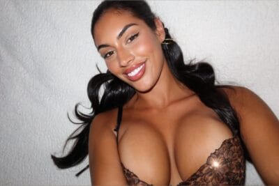 Smiling woman with dark hair wearing black lace lingerie, posing in front of a light textured wall.