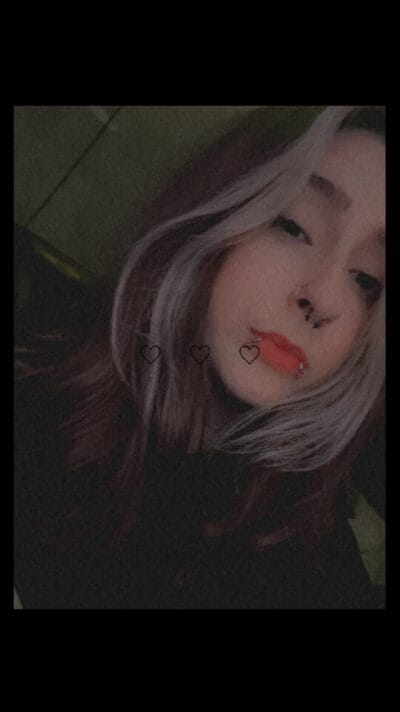 Close-up selfie of a person with grayish hair, bold red lips, and multiple facial piercings; faint heart icons overlay the image.