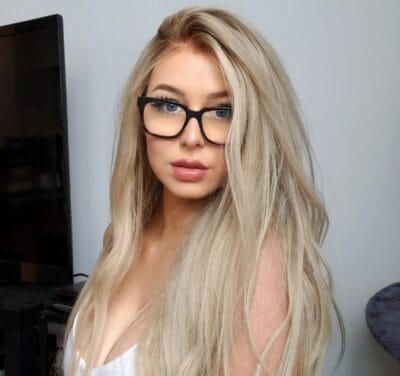 Portrait of a blonde woman wearing large black-framed glasses, posing indoors beside a TV screen on the left.