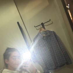 Person taking a mirror selfie with a bright flash; a blue plaid shirt hangs on a rack in the background.