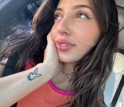 Woman in a car, head resting on hand, looking up, wearing an orange sleeveless top; a bunny tattoo is visible on her forearm with a seatbelt nearby.